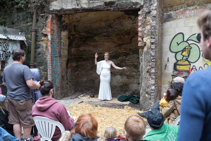 A woman wearing a white costume dress is performing inside a cave, facing outwards to audience