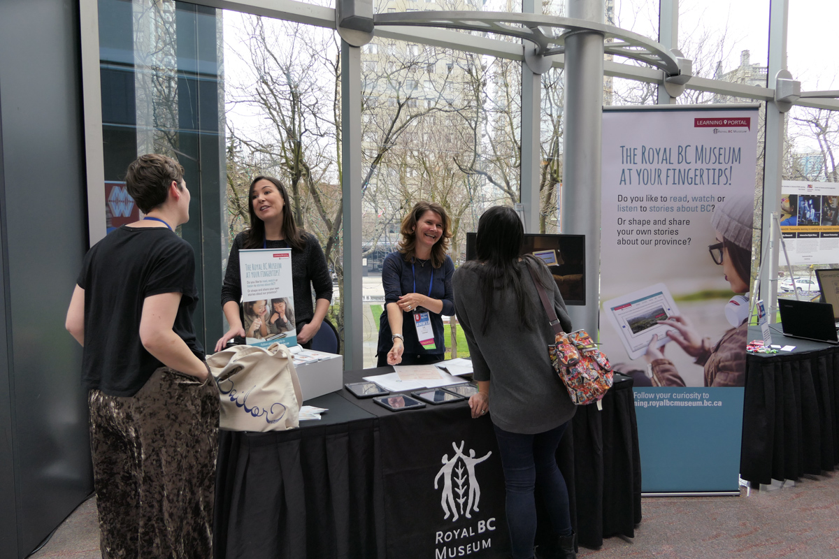 A group of people stand in a bright hallway, talking to exhibitors at MW18.