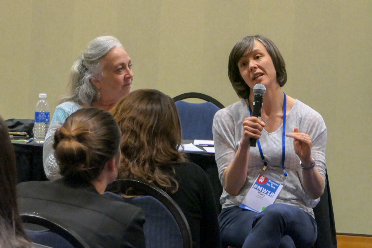 A woman sits in a chair holding a microphone and speaking to a small group of people. 